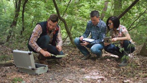 Three casual biologists in woods taking sample of earth in small can holdings digital tab in hands and using silver matal toolbox