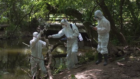 Three bio technicians testing for pollutants in a river in woodland wearing full hazard suits and masks as they take water samples