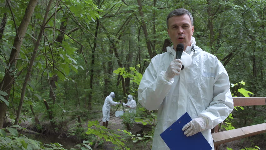 Man wearing protective suit holding file in hands and speaking microphone while looking at camera on nature while other ecologists taking sample of water on background.
