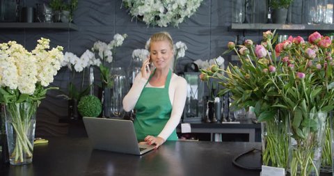 Female florist standing behind counter in shop selling flowers and talking phone while using laptop.