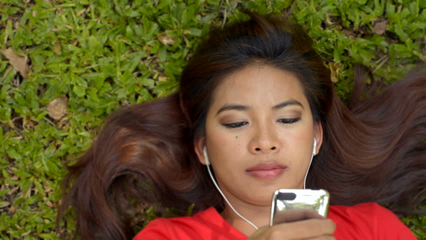 Young Asian woman lying on the grass in a park listening to music on her headphones.