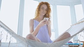 Young positive redhead woman wearing pajamas sitting on hammock and listening music with headphones at home