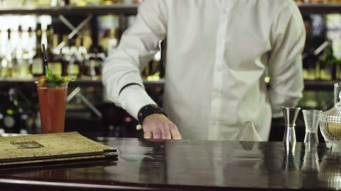 The young barman in white shirt puts a glass with bloody Mary on the coaster next to the menu in the bar slow motion close-up.