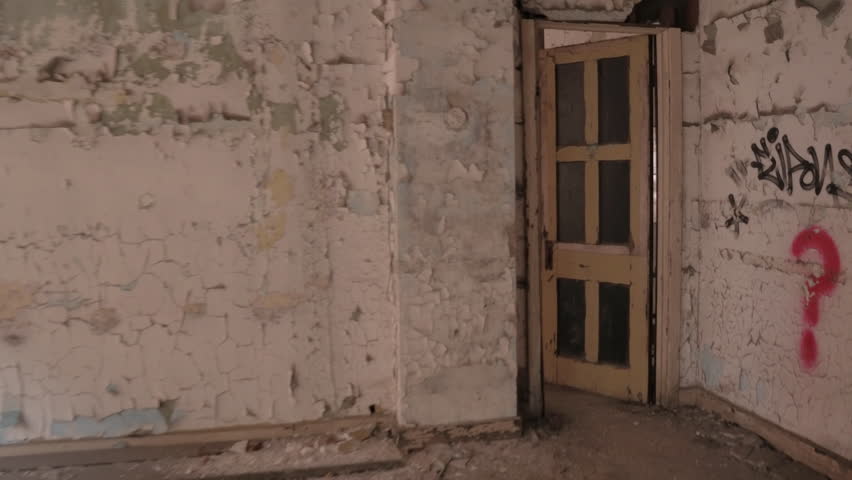 FPV, CLOSE UP: Walking along dark narrow hallway in abandoned crumbling block of flats in decaying ghost town. Weathered wooden doors and peeling paint on the walls in chaotic haunted house in America