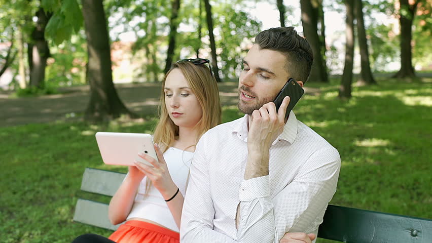 Couple sitting on the bench in the park and using electronics, steadycam shot
