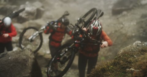 Medium shot of three mountain bikers straight on carrying their bikes up a hill during an extreme enduro race through fog up a mountain. Abstract slow motion shot of extreme sports preparation.
