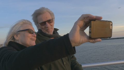 Attractive happy elderly couple in 60s take selfie with smartphone at sunset on ocean smiling and laughing together. Retired baby boomers using cell phone for photos and sharing on social media apps