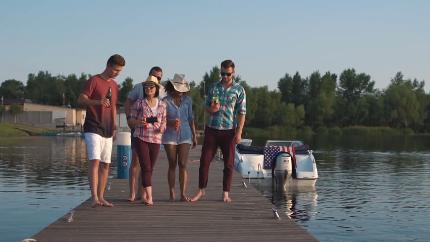 Slow motion of diverse group of young casual people walking on pier with drinks and talking and laugh
