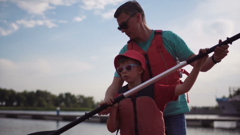 The father teaches the son on pier how to use an oar in a kayak.