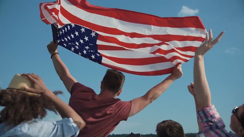 Emotional group of mixed ethnic young friends on board a motor boat along a river laughing and cheering and raise american flag in air. Celebrate an Independence Day on July 4 Stock Video