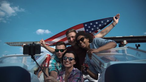 Group of diverse cheering laughing friends on a speedboat waving a patriotic American flag as they pass under a bridge in a low angle view looking up.