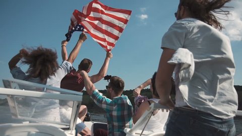 Emotional group of mixed ethnic young friends on board a motor boat along a river laughing and cheering and raise american flag in air. Celebrate an Independence Day on July 4