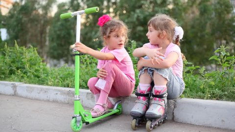 two girls sit on border, one in rollers, another holding scooter, and talk