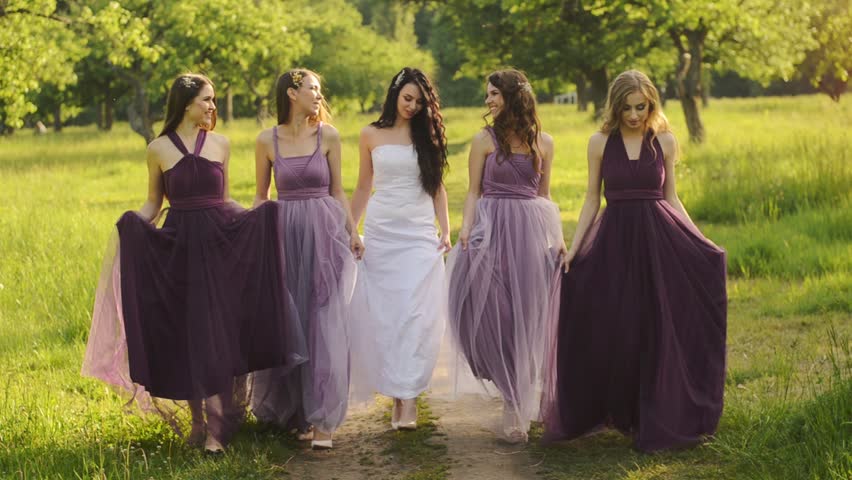 Beautiful and bridemaids in purple dresses walking in the park or garden holding hands and laughing in the evening. Slow motion.