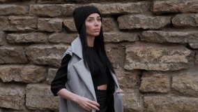 Stylish Fashion Girl, With Long Black Hair, Walking in the Street, on Background Brick Wall, in Grey Cloak and Black Hat