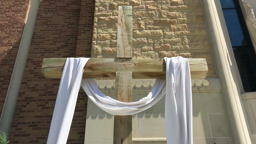 Wooden cross is displayed outside in sunlight with with cloth wrapped around it while wind randomly blows.  