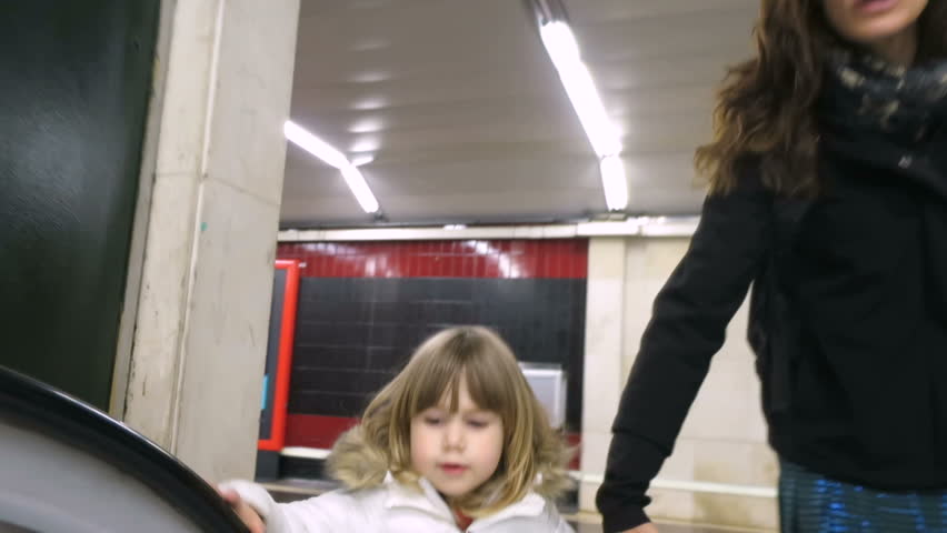 three years old blonde child with white coat holding mother’s hand walking in mechanic stairs of subway or underground station of Madrid city
