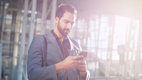 Attractive bearded businessman looking around and using his smartphone while coming out of the modern glassy building, airport or office in a bright light. Stylish look, playful mood.
