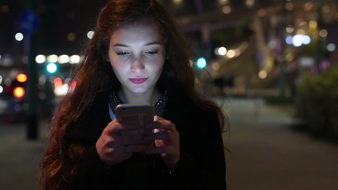 Portrait of young beautiful caucasian woman using smart phone hand hold outdoor in the city night, smiling, face illuminated screenlight - social network, technology, comunication concept