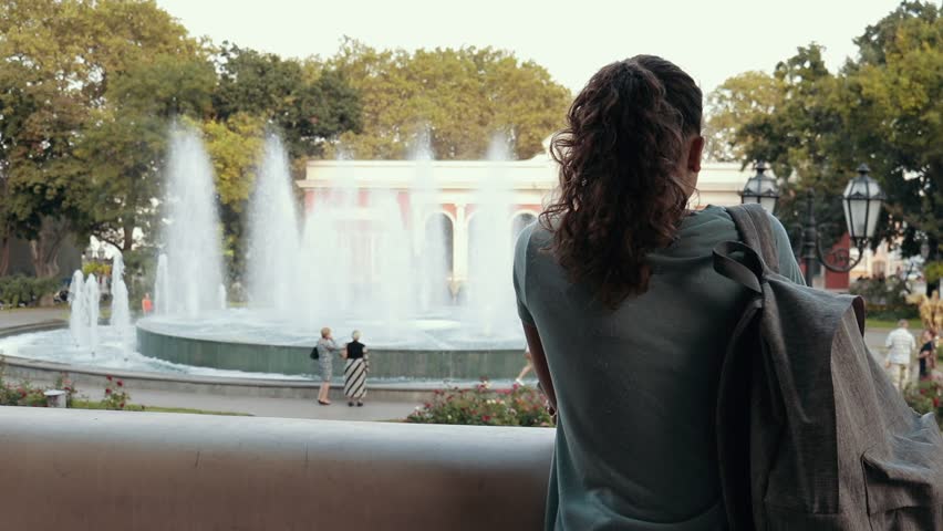 Young woman tourist in a blue t-shirt with a backpack looking at a fountain in an old city in summer. View from the back, slow motion