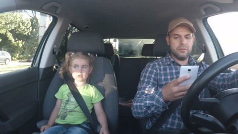 The father and his little daughter are driving in the car. A bad parent uses a smartphone while driving a car. Man texting and driving with his daughter in the car.