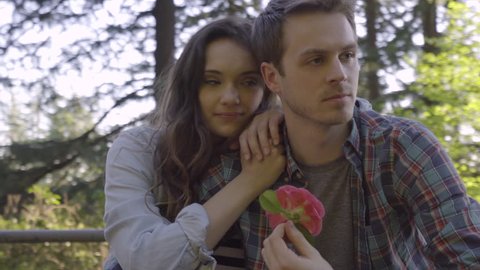 Steadicam Shot Of Couple Relaxing On A Picnic Table Together, Man Twirls A Flower In His Hand, His Girlfriend Snuggles Up To Him