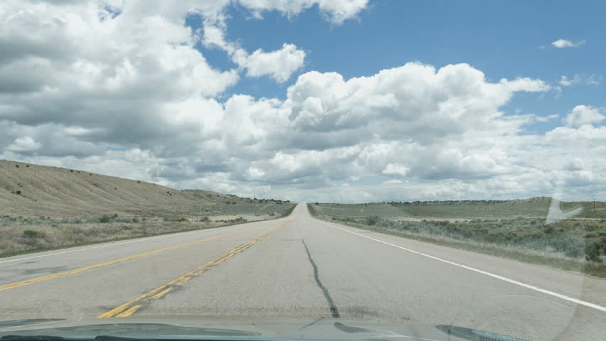 Drive Plate-POV-View forward rolling sagebrush hills western Colorado as road tapers to vanishing point below clouds in a blue sky.