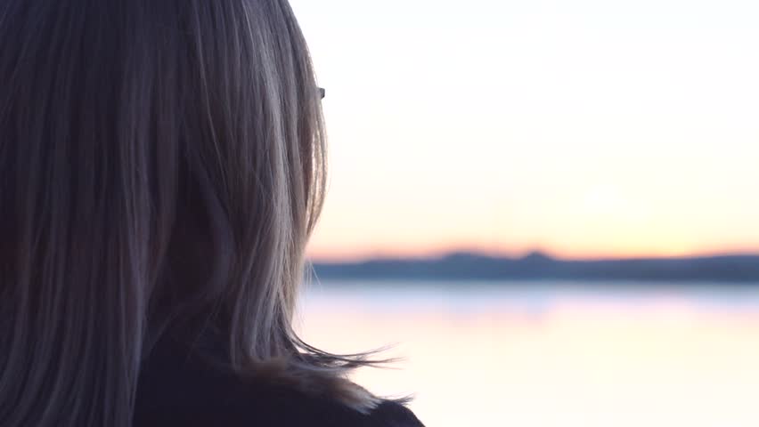 Female in glasses looks at the sunset by the river. Back view