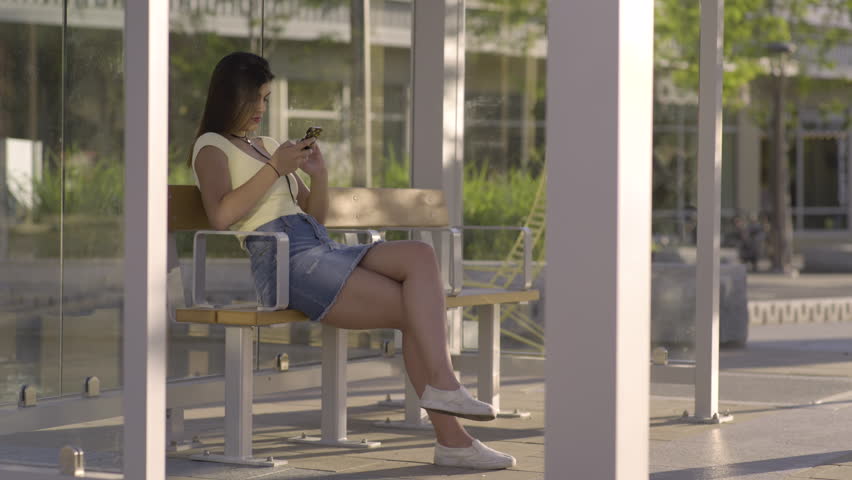 Young Hispanic Woman Plays On Her Smatphone While She Waits For Bus, She Looks Up From Her Phone For Bus