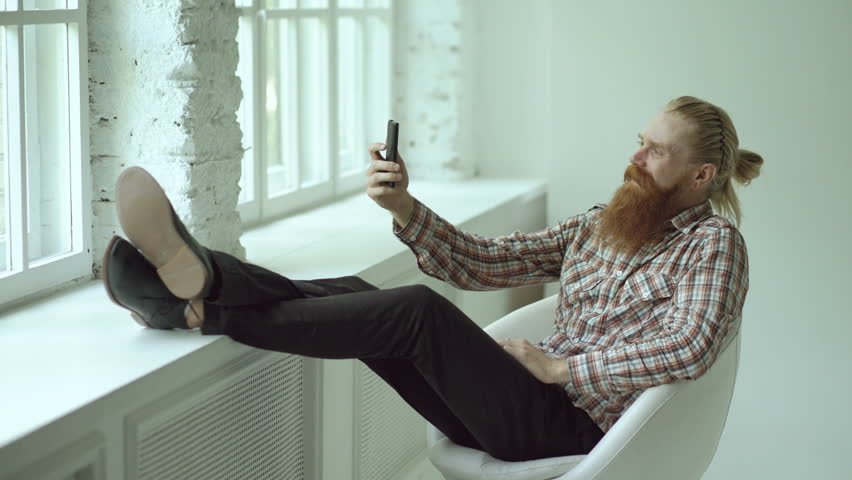 Bearded young hipster businessman talking video chat on smartphone while sitting in offce chair with his legs on windowsill