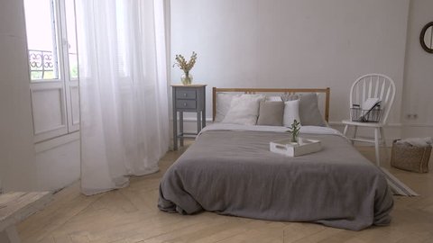 Interior of white and gray cozy bedroom with chair, basket, flowers, bedside table and mirror