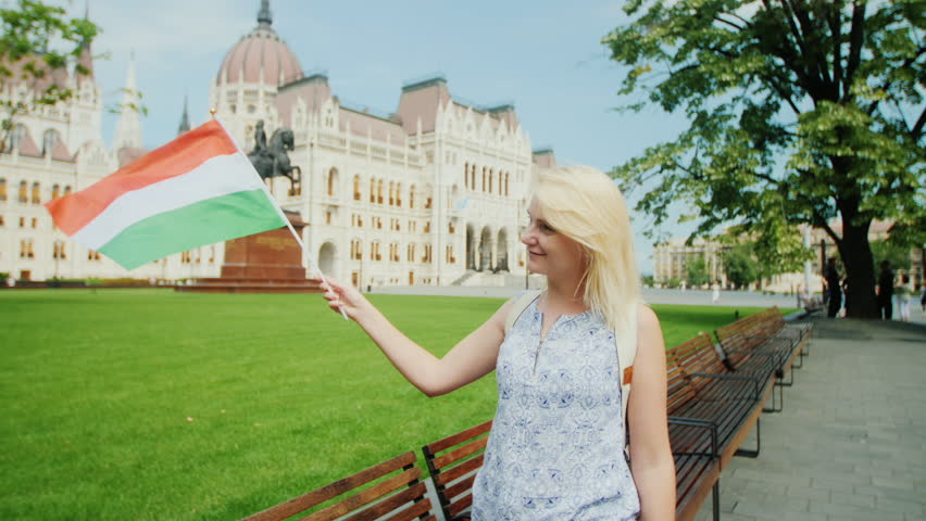 Young woman tourist posing with the Hungarian flag on the background of the parliament in Budapest. Tourism in Europe