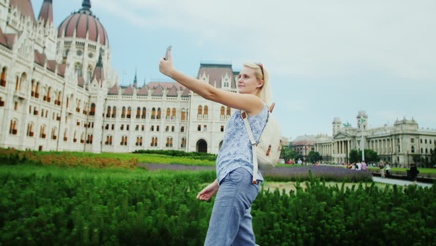 Young woman tourist strolls against the background of the Hungarian Parliament, takes pictures with herself by phone