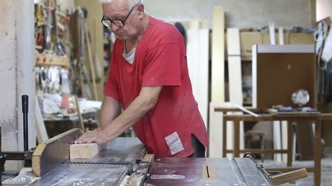 Experienced grey hair carpenter in red shirt wearing glasses doing manual works in his workshop on woodworking machine / Carpenter works in a workshop