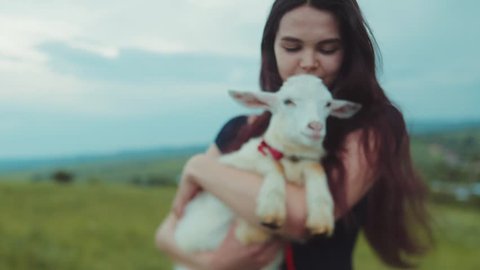 Beautiful long-haired brunette woman holds on her hands white goat kid, kisses it, smiles, pets and strokes it. Loving nature, green peace. Playful mood, being happy. Childhood memories.