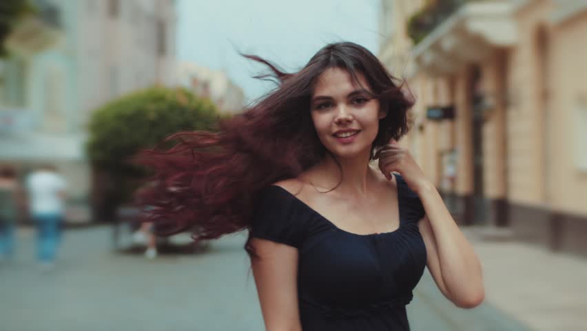 Back view of attractive long-haired curly brunette woman walking down the city-street or alley, turns to camera and gives a beautiful smile, wind plays with her hair. Urban settings on the background.
