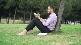 Young, happy businessman watching movie on tablet sitting on grass in park
