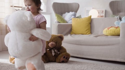 two little happy girl kids play actively with plush toys and teddy bears, hugging them with love and dancing with fun, enjoying and laughing at home in room during sunny day