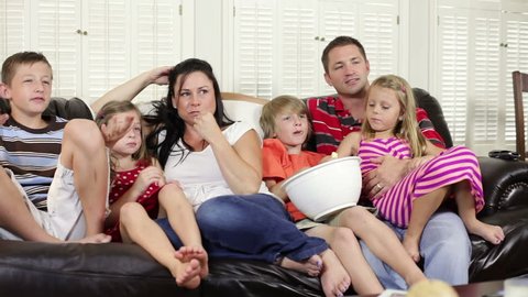 A mom and dad sit on the couch with their four children and watch television. Reveal shot from behind TV.