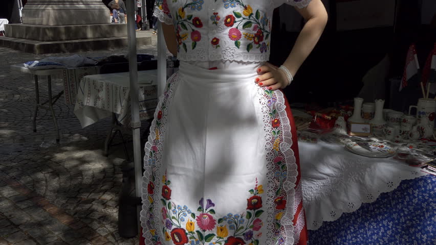Young slim woman wearing national traditional Hungarian clothes and footwear is standing by souvenir stall 