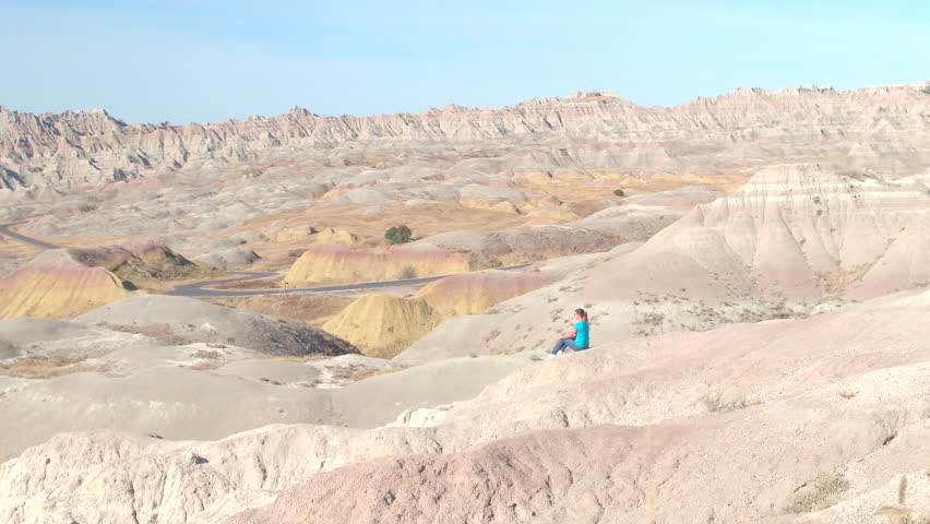 Young woman traveler sitting on rocky mountain slope, admiring the view of stunning Badlands National Park on sunny day in South Dakota, USA. Happy girl exploring sandstone mountain desert wilderness