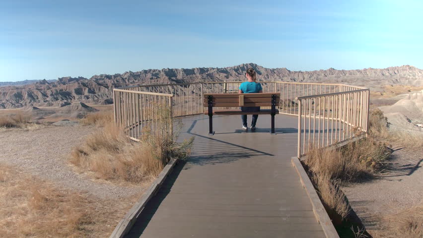 Young woman traveler sitting on bench at the end of a boardwalk, admiring the view of stunning Badlands National Park on sunny day in USA. Happy girl exploring sandstone mountain desert wilderness
