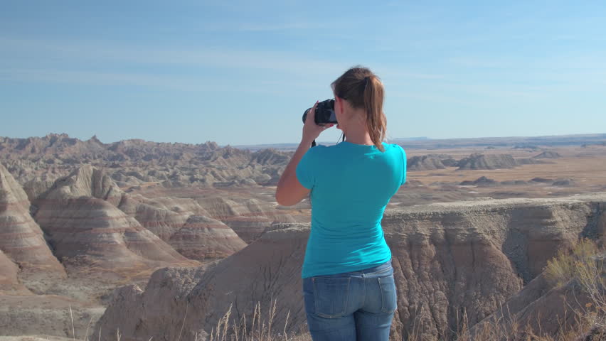 Cheerful young woman traveler taking photos of scenic Badlands National Park on sunny day in South Dakota, USA. Happy girl exploring sandstone mountain desert wilderness and photographing with camera