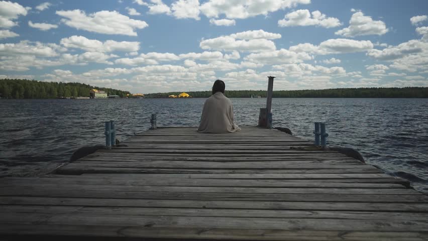 Brunette in plaid sad sitting on an old wooden pier and looks out at the water and forest in the distance.
