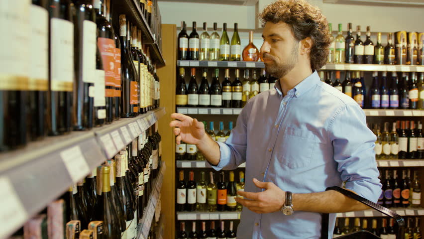 Young attractive man choosing the good bottle of wine in the supermarket and putting it to the basket.
