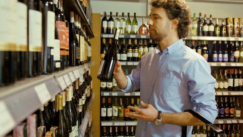 Young attractive man choosing the good bottle of wine in the supermarket and putting it to the basket.