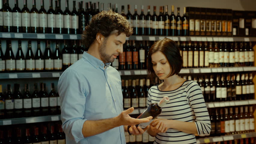 Young attractive couple trying to choose good bottle of wine in the supermarket on background with wine bottle.