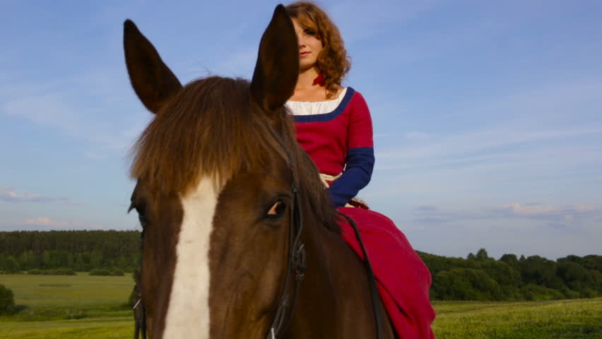 Caucasian girl in vintage long dress sitting on horseback in the evening at sunset looking at the camera. Front view. The horse looks right.hands behind his back, not holding the reins