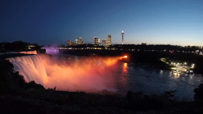 Niagara Falls colorful light illumination from New York State with Canadian skyline. Evening tour boat sailing into mist on Niagara River under fireworks. Time lapse