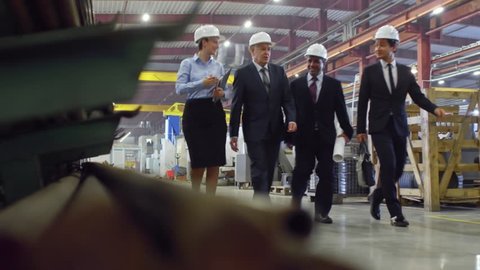 Slow motion shot of team of engineers walking in industrial factory in formal suits and hardhats. Businesswoman telling male colleagues about plant